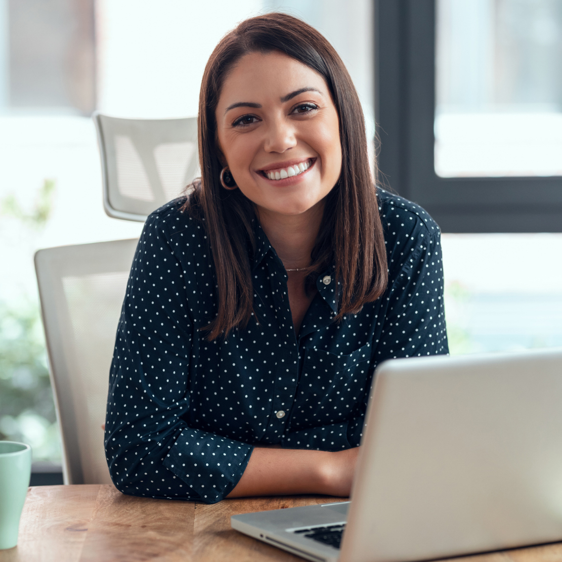 Woman in a polka-dot shirt smiling at a desk beside an open laptop