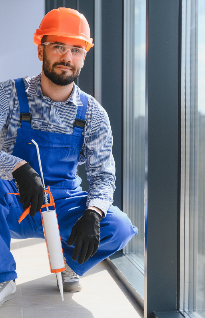Worker in an orange hard hat and blue overalls kneeling with a caulking gun beside a tall window